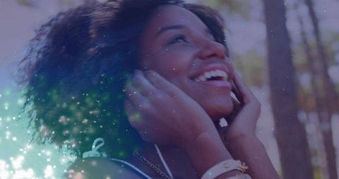 Smiling woman with curly hair listening to music with earbuds in sunlit forest bokeh sunny