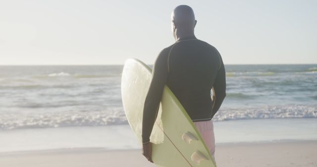 Surfer holding surfboard on the beach while looking at the ocean. Ideal for sports and fitness marketing, travel and adventure advertising, summer vacation promotions, and outdoor lifestyle blogs.
