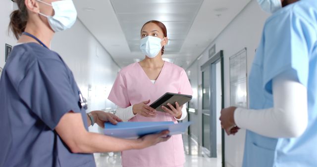 Three healthcare professionals wearing face masks and scrubs are having a discussion in a hospital corridor. One nurse in pink scrubs holds a digital tablet, while the others hold medical documents. This image can be used to depict teamwork, communication, and safety protocols in healthcare settings.