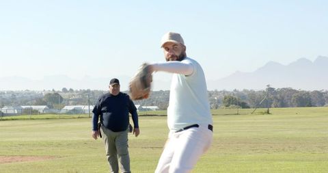 Bearded pitcher throwing from mound with umpire walking behind on sunlit baseball diamond