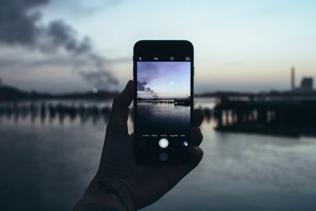 Hand holding smartphone capturing a serene waterfront during sunset. Perfect for promoting mobile photography, peaceful scenes, and serenity in urban nature. Can be used in articles about the progression of technology in photography and nature-related themes.