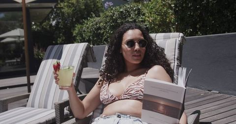 Reclining woman sipping cocktail and reading paperback on striped pool lounger