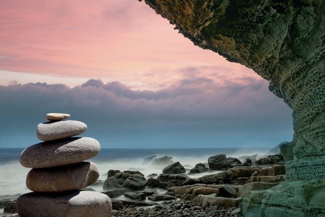 Serene coastal scene featuring a neat stack of stones with rocky cliffs and calming ocean waves at sunset. Captures tranquil and meditative atmosphere perfect for mindfulness, zen themes, and nature-related content.