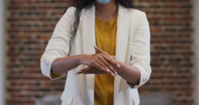 Businesswoman standing against a brick wall, demonstrating hand sanitizing technique while wearing a face mask. Emphasizing hygiene and workplace safety during pandemic, makes this suitable for articles or visuals related to COVID-19 safety measures, workplace health guidelines, or office hygiene practices.