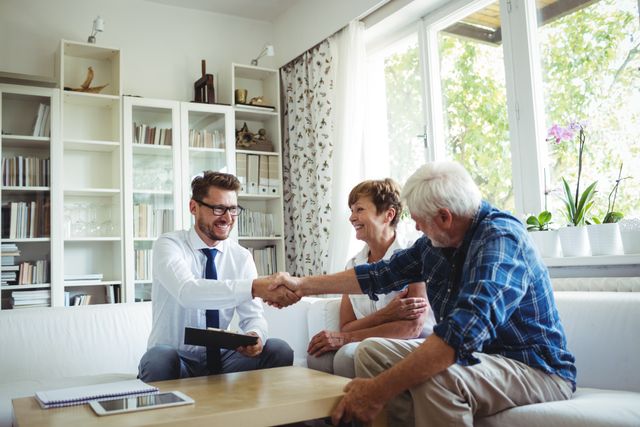 Financial advisor shaking hands with senior man while senior woman watches in living room. Ideal for use in articles or advertisements about financial planning, retirement planning, trust in financial advisors, and professional consultations. Can be used to depict positive client-advisor relationships and home consultations.