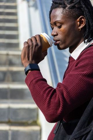 African american man sipping coffee on city steps wearing maroon sweater, smartwatch