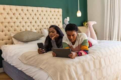 Diverse women relaxing on bed using tablet and smartphone, cozy modern bedroom lifestyle