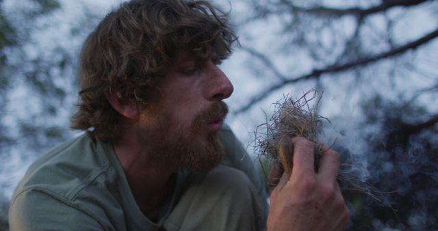 This photo shows a man blowing into dried grass to start a campfire in a wilderness setting. Perfect for articles and blogs on outdoor survival skills, camping tips, or nature documentaries. Ideal for use in advertisements or promotional materials for outdoor gear and adventure experiences.