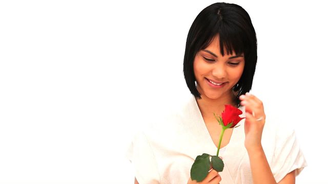 This image portrays a woman smiling gently while holding a red rose. The white background emphasizes the rose's vibrant color and the woman's serene expression. This photo is ideal for use in articles about love, beauty, or special occasions, and it can also be used in promotional materials or romantic greeting cards.