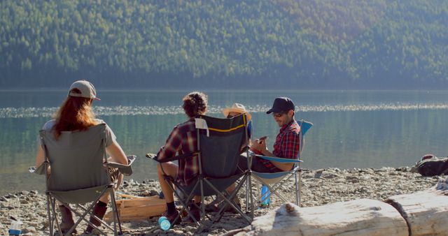 Group of friends sitting on camping chairs next to tranquil mountain lake, enjoying conversation. Ideal for depicting lifestyle moments, summer adventures, travel destinations, outdoor relaxation, friendship, and leisure time in nature settings.