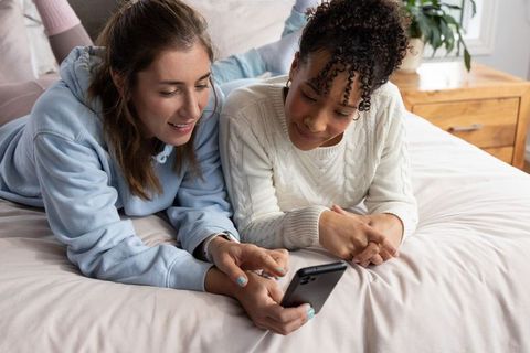 Multiracial friends relaxing on bed sharing smartphone content in cozy pastel bedroom