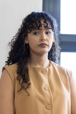 Young woman in tan sleeveless top standing by window with gold hoop earrings, minimalist portrait