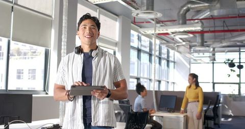 Smiling young professional holding tablet with headphones in modern open-plan office