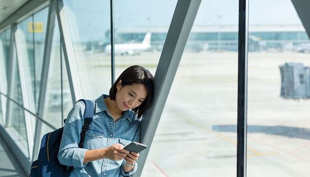 Young woman checking phone in airport walkway leaning on diagonal column with backpack