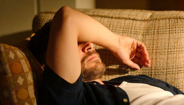 Man resting on sofa shielding face with arm in warm evening light conveying fatigue and quiet recove