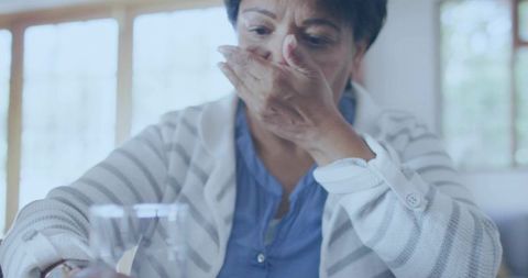 Senior indian woman covering mouth while leaning at table with glass in bright living room