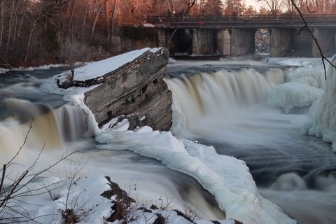 Winter waterfall cascading past snow-capped rock with icy river and bridge at dusk