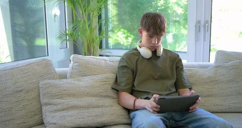 Teen boy relaxing on sofa using tablet with headphones in bright living room