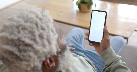 Senior african american man reclining on couch using smartphone with blank screen mockup