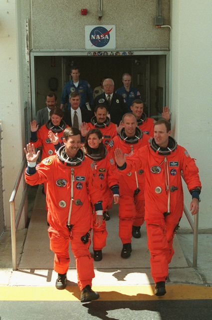 Group of astronauts are seen waiving to onlookers as they head from the Operations and Checkout Building for their third launch attempt on Space Shuttle Atlantis. The image includes Pilot Scott J. Horowitz, Commander James D. Halsell Jr., and Mission Specialists Mary Ellen Weber, Jeffrey N. Williams, Susan J. Helms, Yury Usachev, and James S. Voss in orange spacesuits. The mission involves logistics delivery to the International Space Station, preparations for the Russian Zvezda Service Module, and a spacewalk. Suitable for illustrating topics related to NASA, space exploration, teamwork, and space shuttle missions.