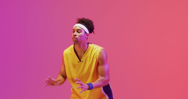 This image depicts a young black man playing basketball in an indoor studio with a vibrant colored background. He is wearing a yellow jersey and a white headband, demonstrating focus and intensity. Useful for marketing campaigns, sports-related advertisements, athletic product promotions, fitness blogs, and motivational posters. Ideal for illustrating themes of athleticism, energy, and dedication.