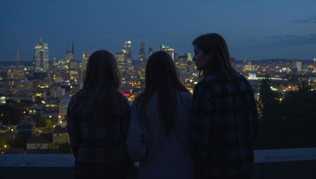 Three friends standing on rooftop railing at dusk gazing over illuminated city skyline