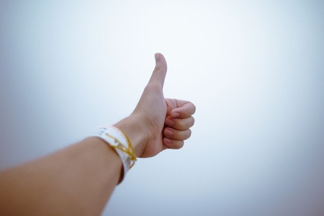 Hand showing thumbs up gesture, symbolizing approval, agreement, or positivity. Person wearing a wristband with multi-colored design. Suitable for use in positive messages, success stories, approval, and marketing materials.