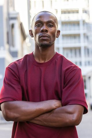 African american man standing with crossed arms on city street wearing maroon shirt