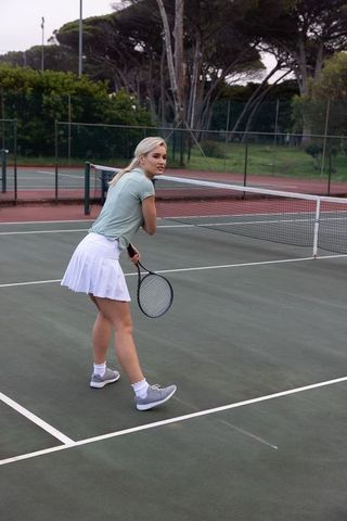 Young woman practicing tennis at net wearing green top and white skirt on outdoor court