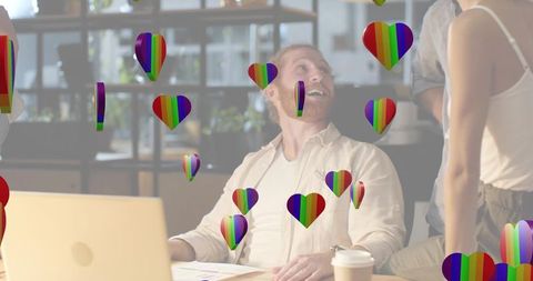 Laughing man collaborating at laptop surrounded by rainbow hearts in startup office