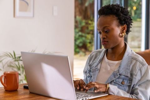 African american woman working on laptop at home in bright casual workspace with coffee mug