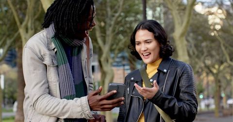 Diverse friends sharing phone and laughing in urban park during autumn stroll