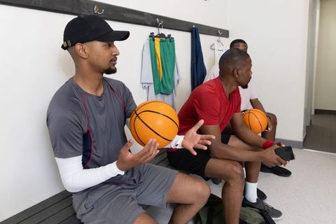 Diverse basketball teammates sitting on bench holding balls, using phone and socializing before game