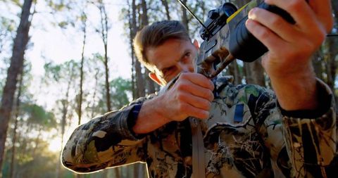 Mature hunter aiming crossbow in sunlit pine forest wearing camouflage jacket