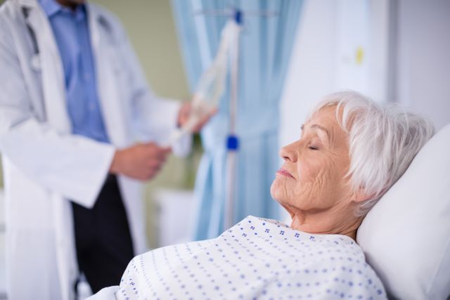 Senior woman resting in a hospital bed with a doctor in the background holding an IV drip. Ideal for use in healthcare, medical treatment, elderly care, and hospital-related content. Can be used in articles, brochures, and websites focusing on patient care, recovery, and medical services.
