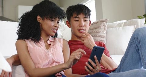 Diverse young couple relaxing on sofa sharing smartphone at home