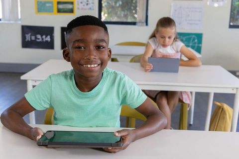 Smiling student holding tablet in classroom while classmate using tablet and math posters