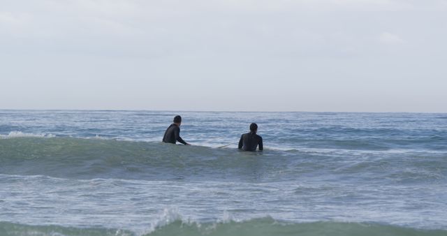 Two surfers wearing black wetsuits waiting on their boards in calm ocean waters. Ocean horizon is visible in the background. Ideal for use in content about surfing, water sports, ocean scenes, calm and serene nature, or sportswear advertisements.