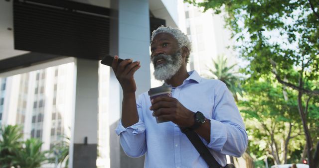 Older businessman with white hair and beard in modern urban area, holding coffee and using smartphone for voice communication. Suitable for themes related to business, corporate life, urban living, professional lifestyle, technology use in daily life, and the blend of work and relaxation. Ideal for advertising tech products, business solutions, corporate environments, and urban development.