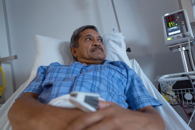 Mature male patient lying in hospital bed, undergoing medical treatment. Hospital monitor and medical equipment visible. Ideal for use in healthcare articles, medical care brochures, patient recovery stories, and hospital website imagery.