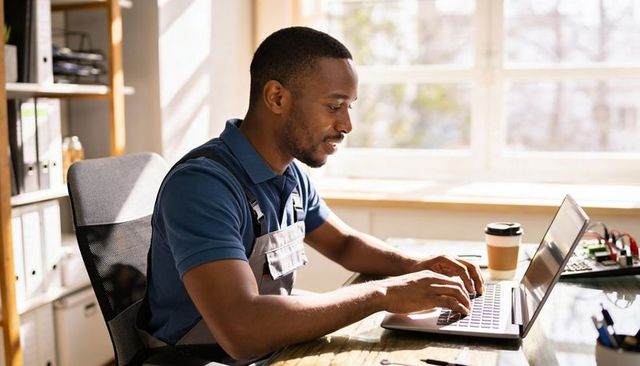 Male technician working on laptop at sunlit workshop desk with tools and coffee cup