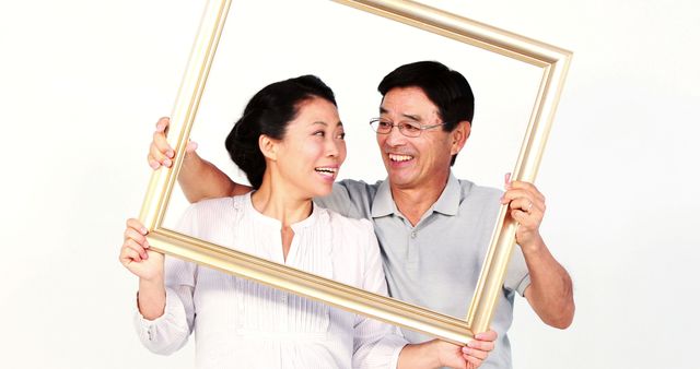 A middle-aged Asian couple shares a joyful moment holding a large picture frame, with copy space. Their smiles and playful interaction suggest a warm, loving relationship.