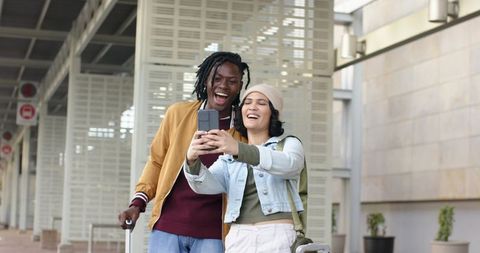 Multicultural couple taking selfie in modern transit concourse holding smartphone and rolling luggag