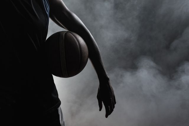 This image captures a closeup of a man holding a basketball in dark lighting, creating a dramatic and moody atmosphere. The silhouette effect and smoke add intensity, making it suitable for use in sports promotions, motivational posters, and advertisements targeting athletes or sports enthusiasts.