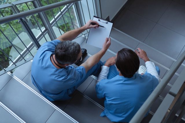 Two male surgeons in blue scrubs are sitting on hospital stairs, engaged in a discussion over a clipboard. This image can be used to depict teamwork and collaboration in a healthcare setting, medical consultations, or the daily life of medical professionals. Ideal for use in medical articles, healthcare blogs, hospital websites, and educational materials.