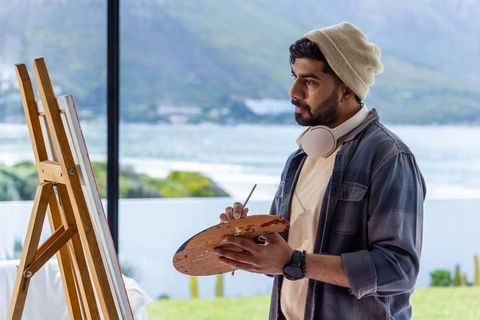 Indian artist painting coastal landscape on canvas with palette and headphones