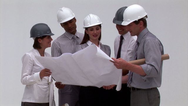 Groups of architects in hard hats examine blueprint together. The setting emphasizes teamwork and collaboration in a construction or architectural workforce environment. Useful for conveying concepts of planning, engineering expertise, diversity in the field, and business strategy in construction materials development.