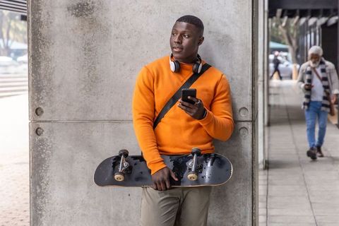 African american skateboarder leaning on pillar in orange sweatshirt holding phone
