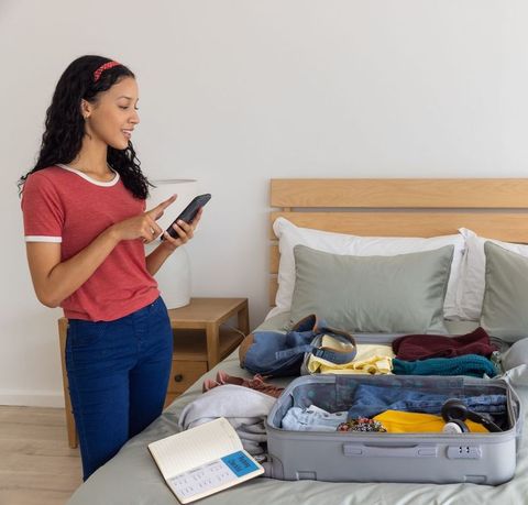 Teenage girl packing suitcase while checking smartphone in minimalist bedroom