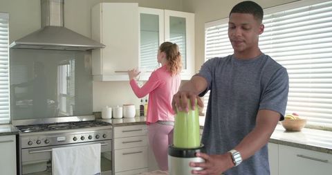 Diverse couple making green smoothie in bright modern kitchen with stainless appliances